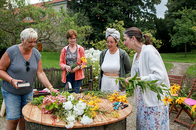 Vier Frauen stehen um eine runde Holzplatte, auf der Blumen liegen.